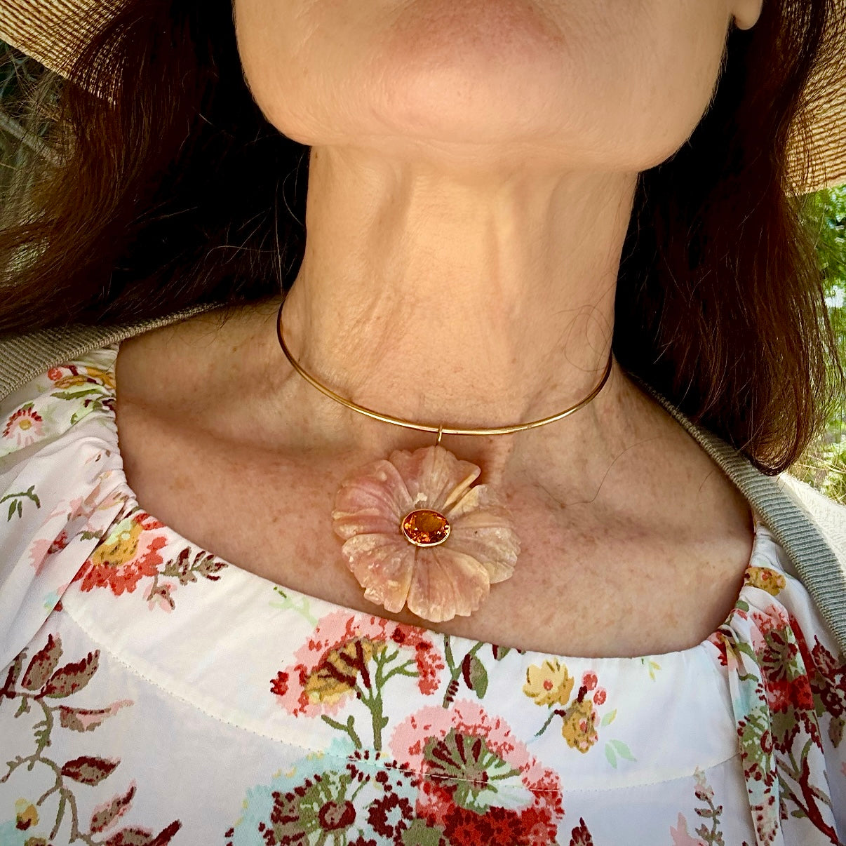 A close-up lifestyle shot of a woman wearing a delicate gold choker necklace with a large, textured pale pink flower pendant featuring a central amber-orange stone.