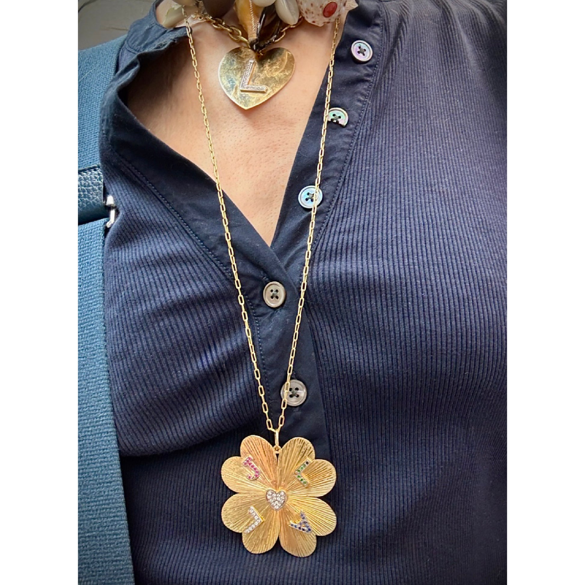 A gold flower-shaped pendant necklace with colorful enamel details and a small heart pendant, worn on a model's neck over a navy blue button-up shirt.