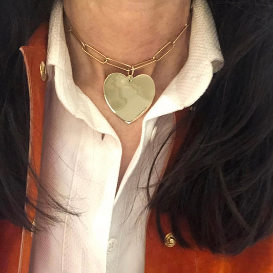 Person wearing a gold necklace with a heart-shaped pendant, white shirt, and brown jacket.