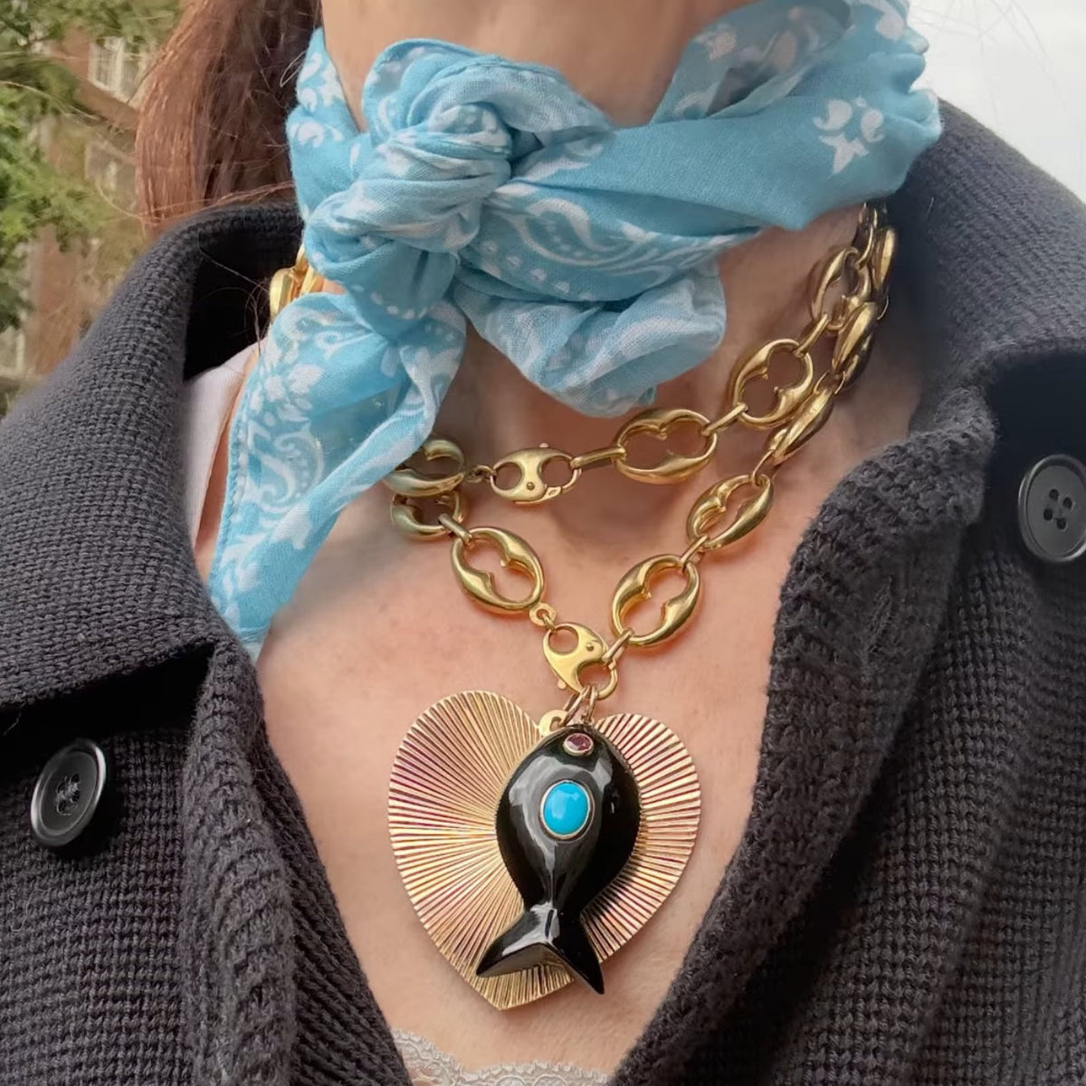 A close-up of a person wearing a gold chain necklace with a heart-shaped sunburst pendant and a black fish charm featuring a turquoise center. The person is also wearing a light blue patterned scarf and a dark gray knit cardigan.