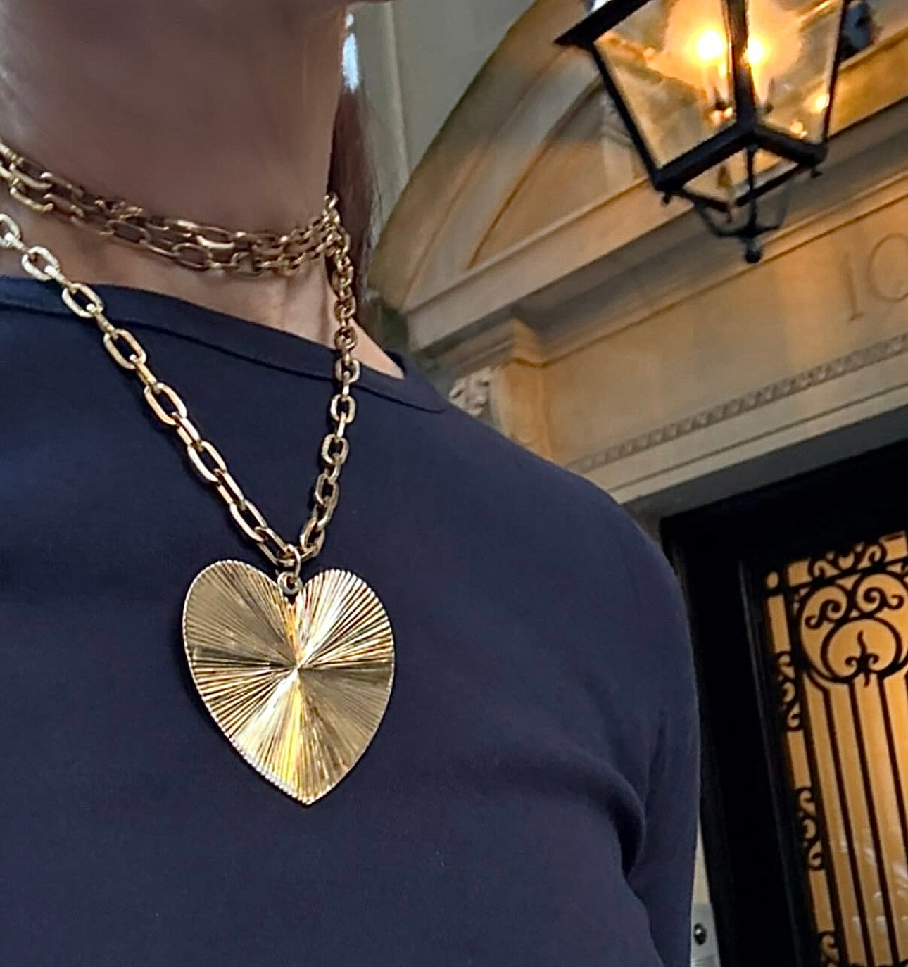 Person wearing a gold heart-shaped necklace in front of a building entrance.
