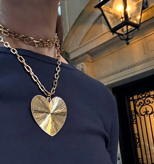 Person wearing a gold heart-shaped necklace in front of a building entrance.