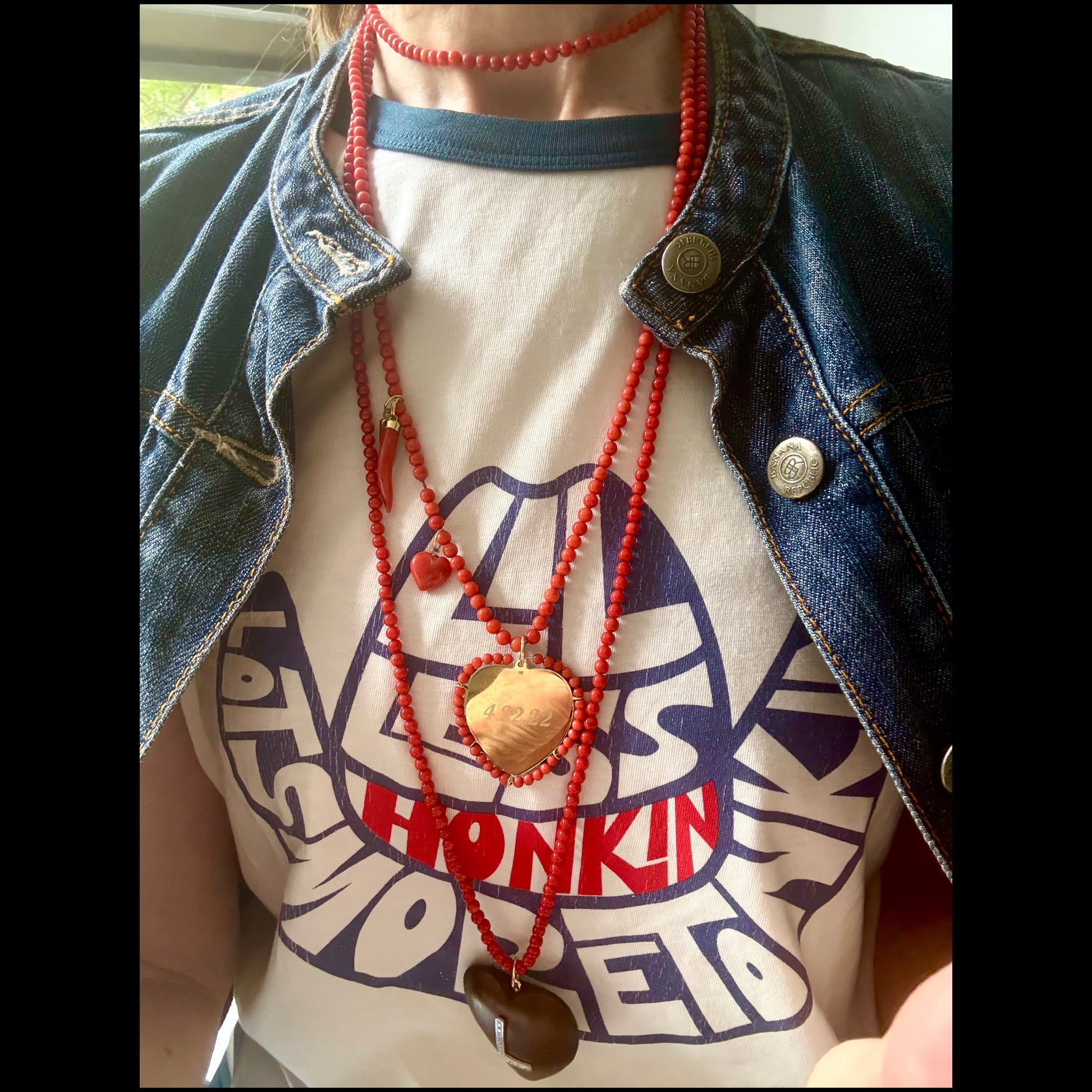 A person wearing layered red coral beaded necklaces with wooden and gold heart pendants over a white graphic t-shirt and an open blue denim jacket.