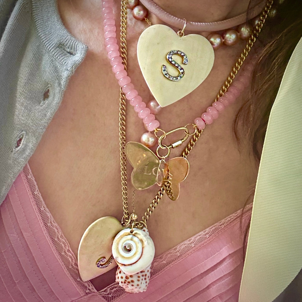 A close-up of a person wearing layered necklaces over a pink lace top and gray cardigan. The necklaces include a large gold heart pendant with an 'S' charm, pink beaded strands, gold chain links, butterfly charms, shell charms, and pearl accents.