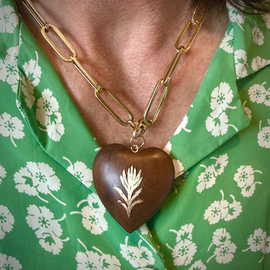 A close-up of a person wearing a gold paperclip chain necklace with a dark brown wooden heart pendant featuring a white floral engraving, paired with a green and white floral shirt.