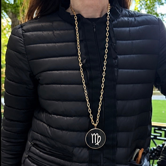 A gold chain necklace with a circular pendant featuring the Virgo zodiac symbol, worn by a person in a black puffer jacket outdoors.