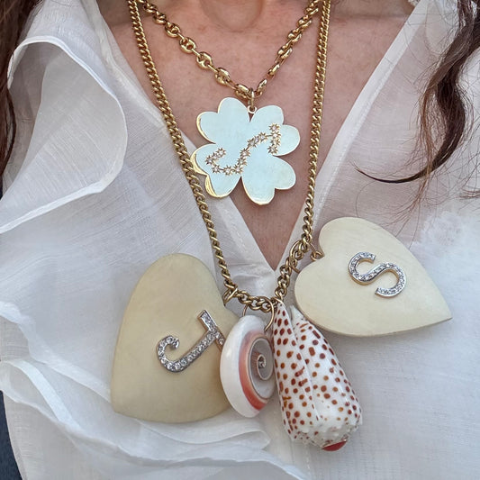 A close-up of a layered necklace worn on a model's neck. The necklace features a gold chain with a large clover pendant, a gold chain with two cream-colored heart pendants (one with a diamond 'S' and one with a 'J'), a small white and coral seashell, and a larger patterned seashell with brown spots.