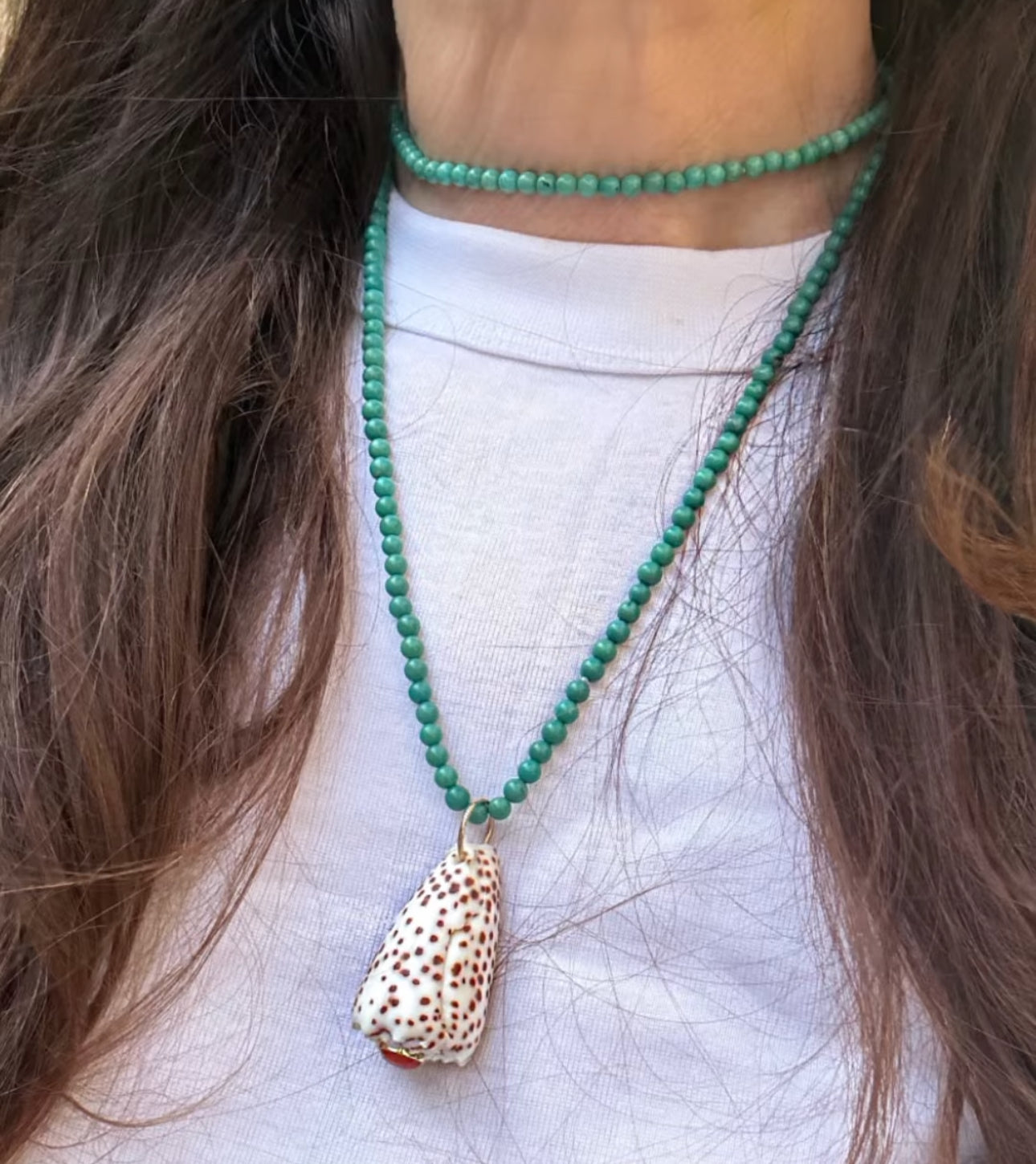 A close-up of a person wearing a green beaded necklace with a Leopard Cone shell pendant, layered over a turquoise beaded necklace, worn with a white t-shirt.