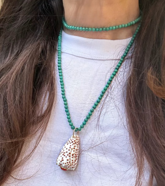 A close-up of a person wearing a green beaded necklace with a Leopard Cone shell pendant, layered over a turquoise beaded necklace, worn with a white t-shirt.