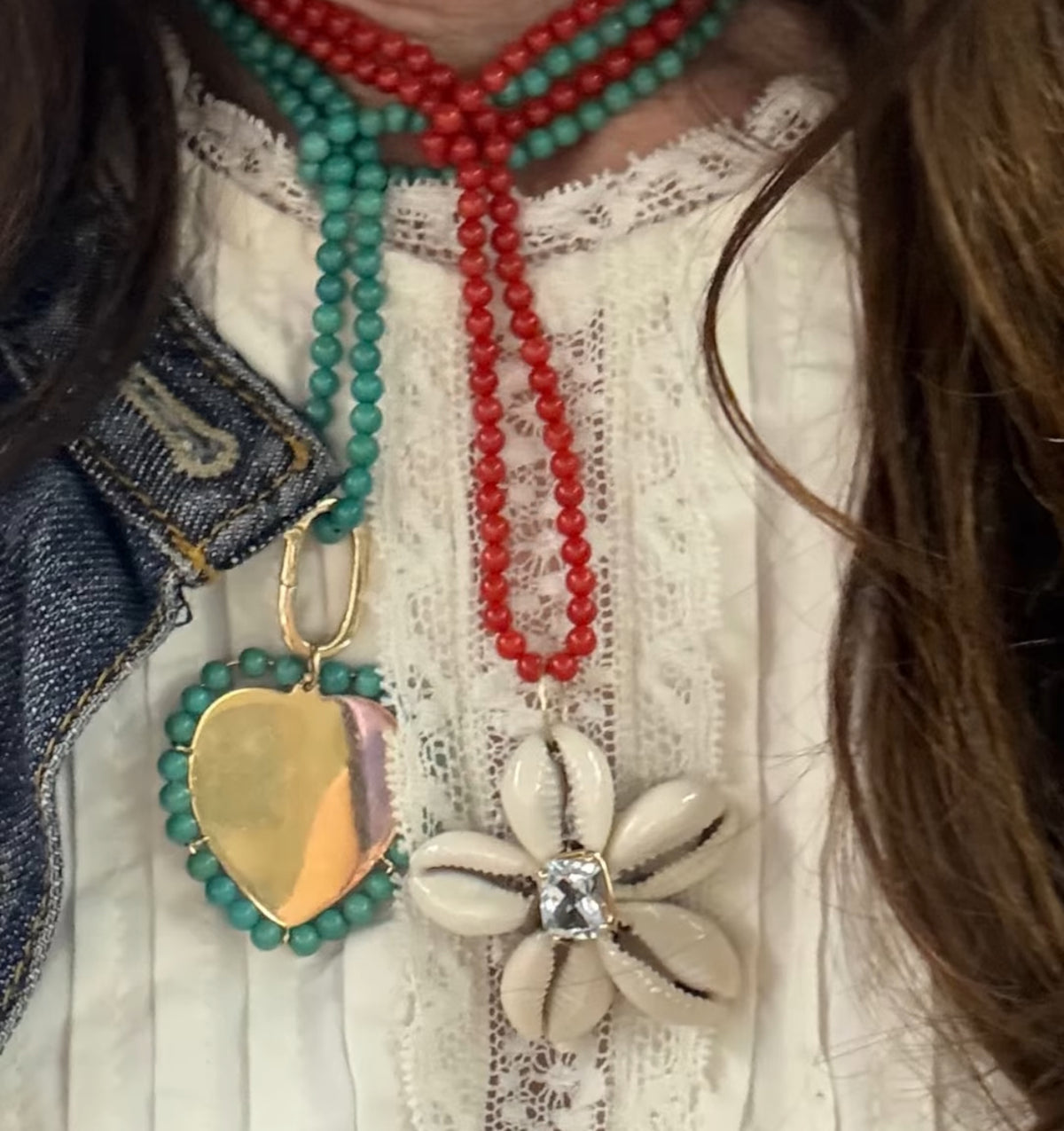 A close-up of a person wearing layered beaded necklaces over a white lace top and denim jacket. One necklace has turquoise beads with a gold heart-shaped pendant, another has red coral beads, and a third necklace features cream-colored cowrie shell flower pendants with a clear rhinestone center.