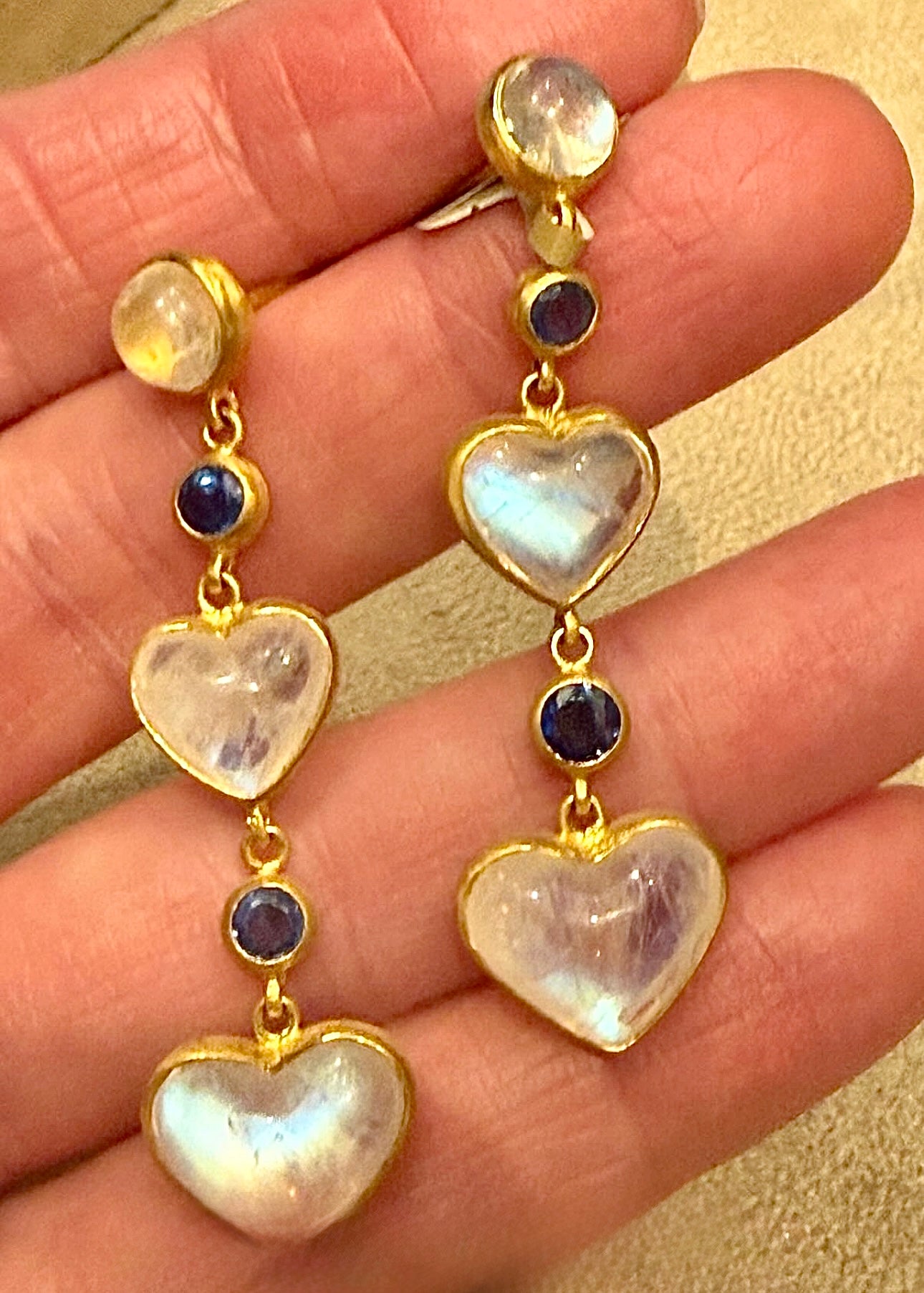 Heart-shaped earrings with gold and gemstone details held in a hand against a neutral background