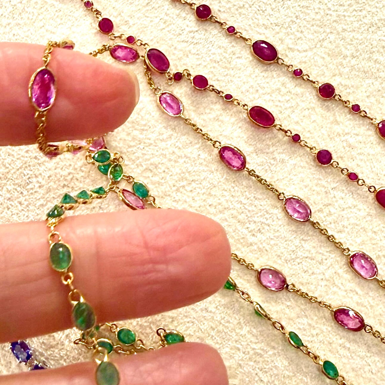 Gold chain necklace with pink and green gemstones held by a hand on a beige background