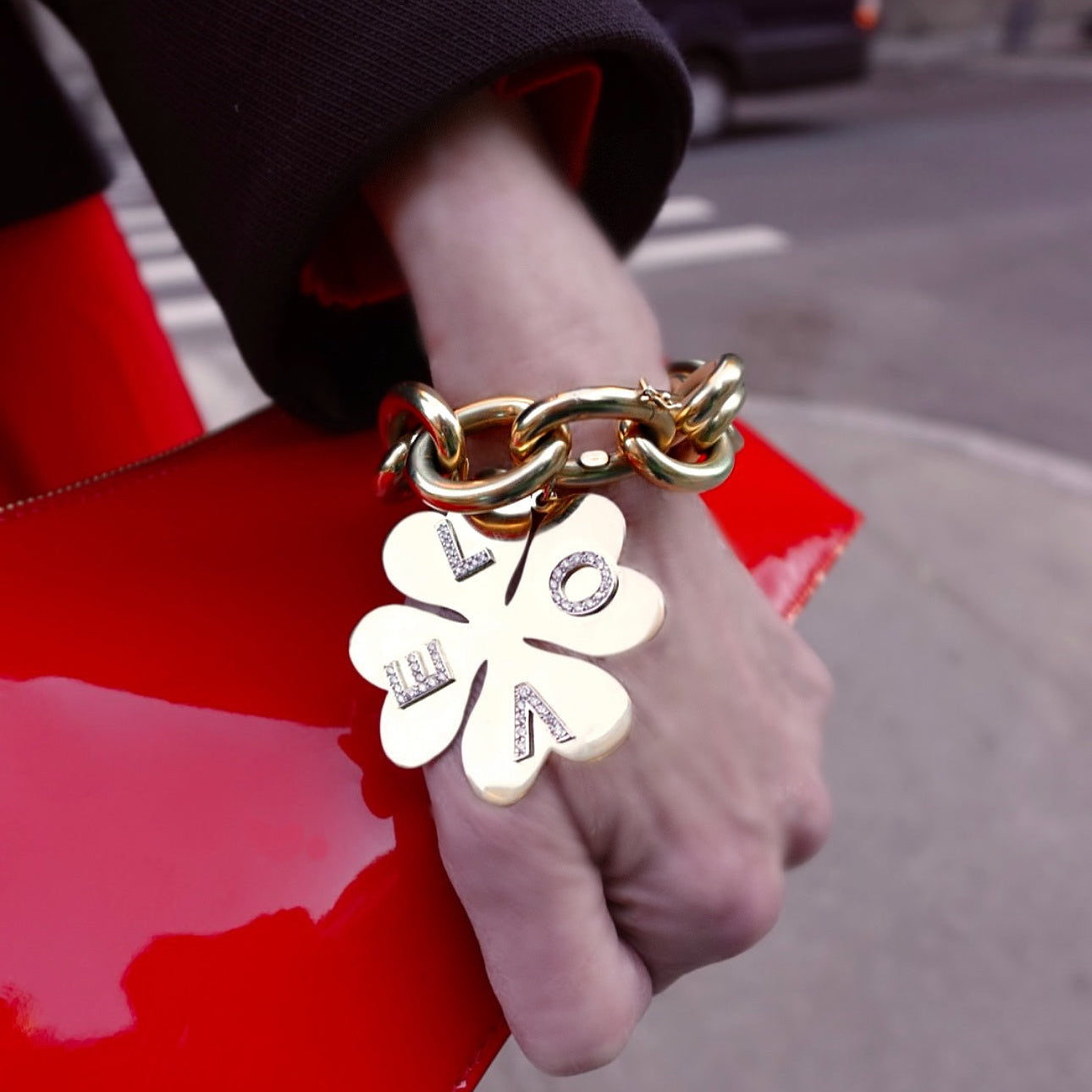 Gold chain bracelet with a clover-shaped charm held by a hand on a red surface.