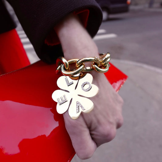 Gold chain bracelet with a clover-shaped charm held by a hand on a red surface.