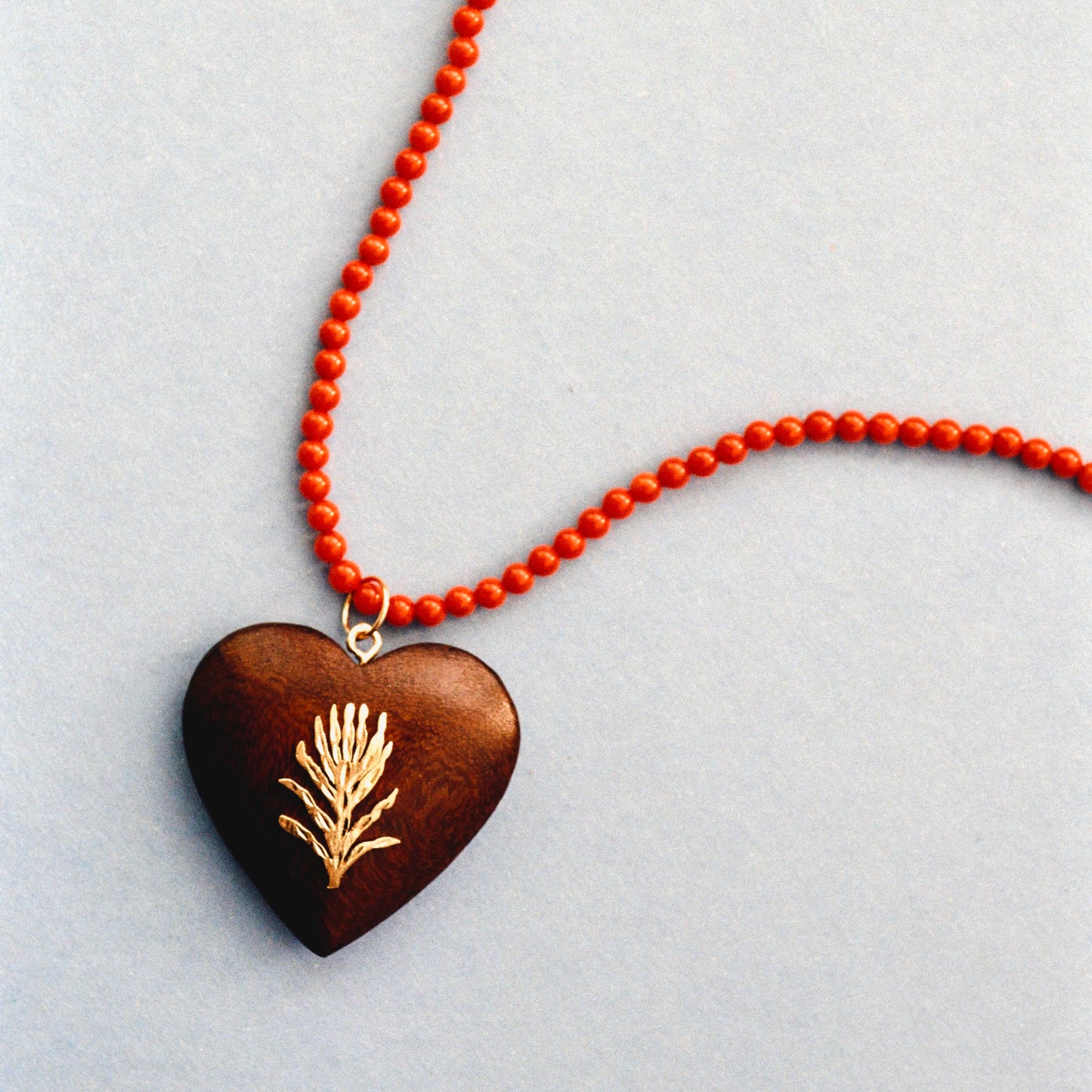 A heart-shaped wooden pendant necklace with a gold pine branch engraving, suspended from a bright orange-red beaded chain on a light gray background.