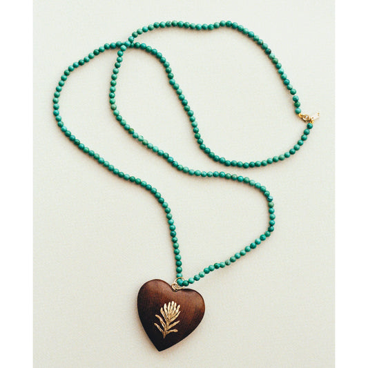 A beaded necklace with vibrant turquoise green beads and a dark brown wooden heart pendant featuring a gold leaf design, displayed on a light beige background.