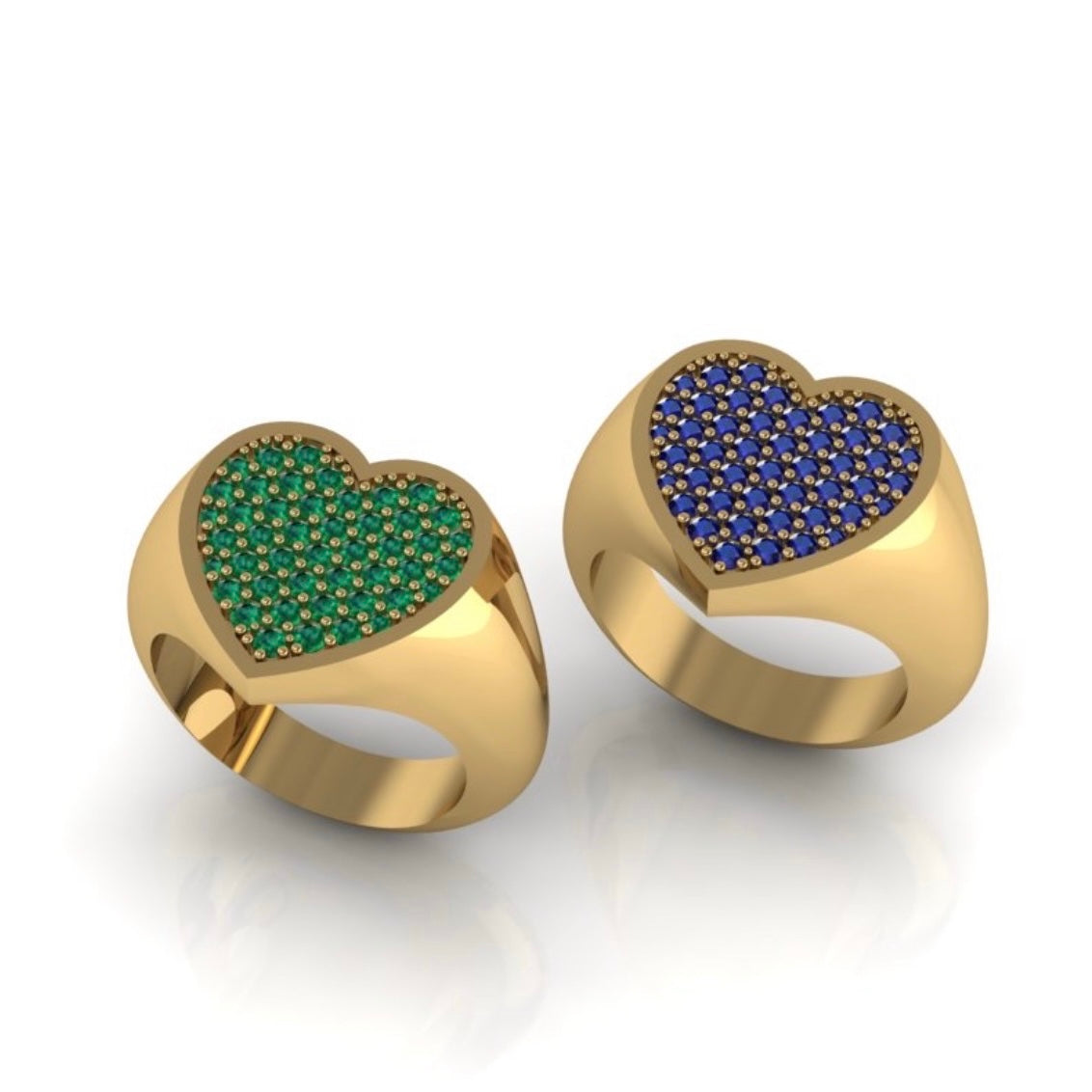 Two heart-shaped gold signet rings on a white reflective background. One ring features a green pavé heart face, and the other features a blue pavé heart face.