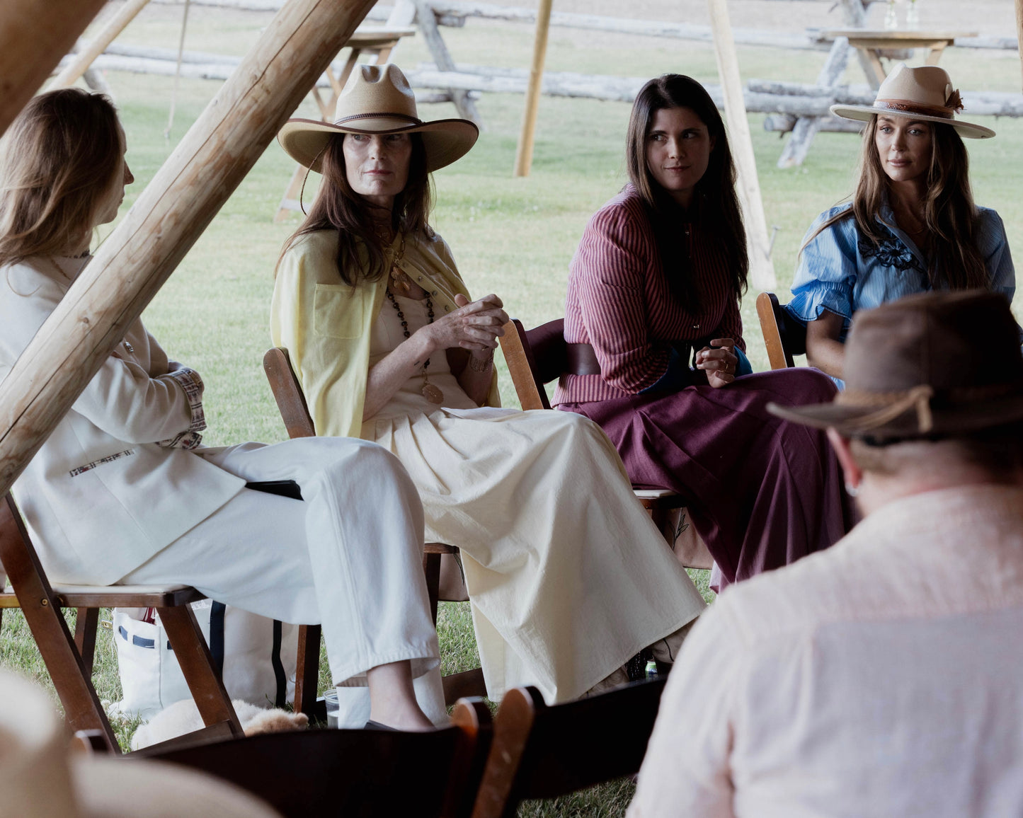Four women seated outdoors under a wooden structure, wearing wide-brim hats and casual western-inspired clothing in cream, yellow, burgundy, and blue.