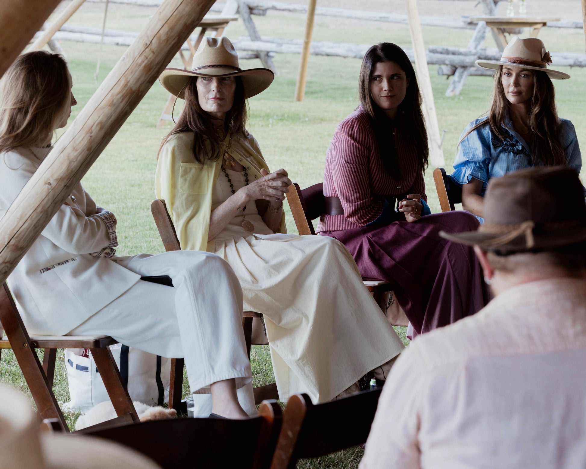 Four women seated outdoors under a wooden structure, wearing wide-brim hats and casual western-inspired clothing in cream, yellow, burgundy, and blue.